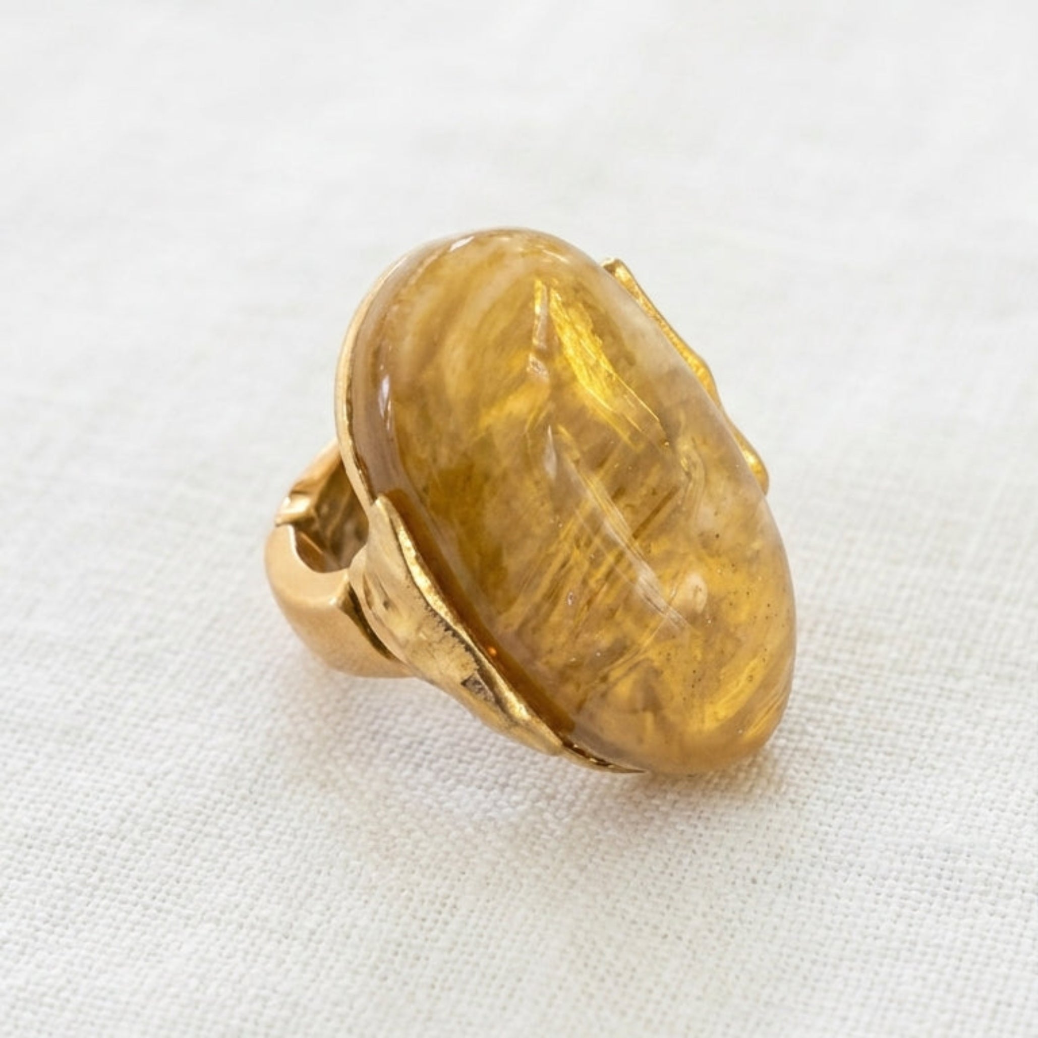 Envy gold ring featuring a large, polished oval cabochon gemstone with a warm amber-yellow hue, resting on a textured white fabric surface.