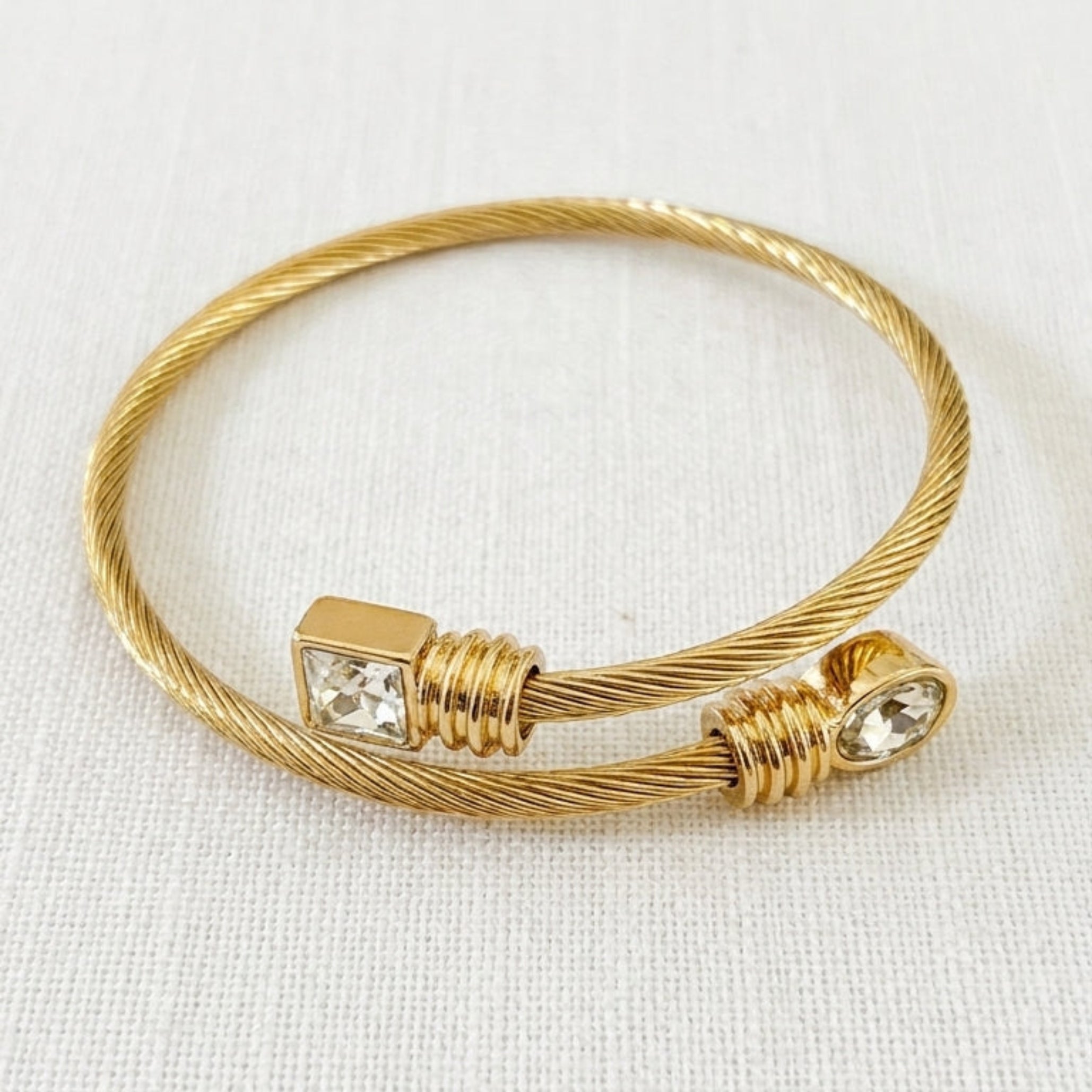 Envy gold twisted cable open bangle with square crystal and round crystal ends on cream linen background