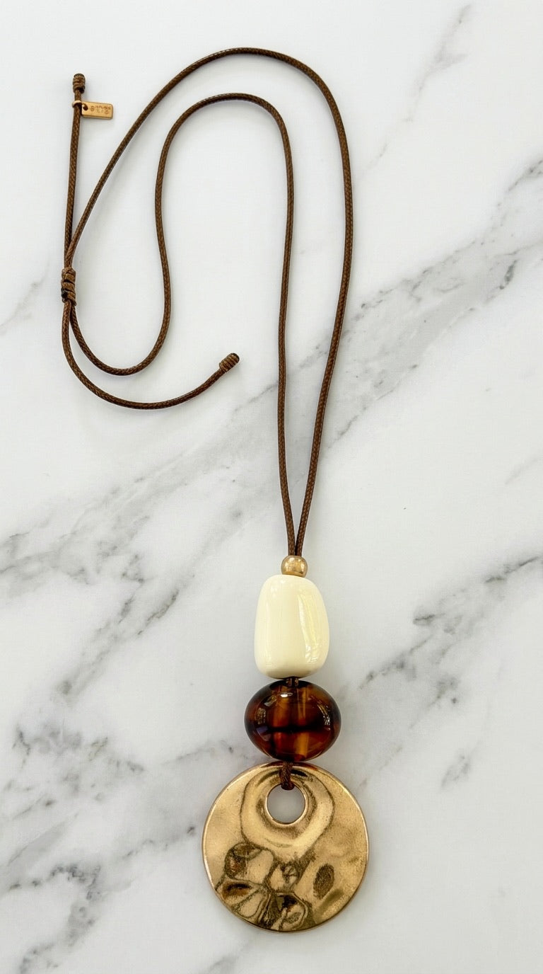 Envy long brown cord necklace with stacked cream resin, tortoiseshell disc, and hammered gold coin pendant on marble background