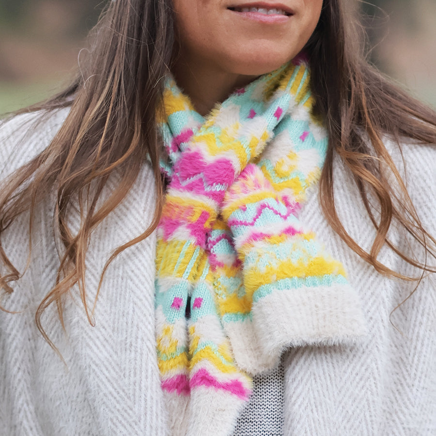 Model wearing the POM Mia pull-through scarf in cream, pink, yellow and mint fair isle pattern, styled through a white chunky knit cardigan outdoors.