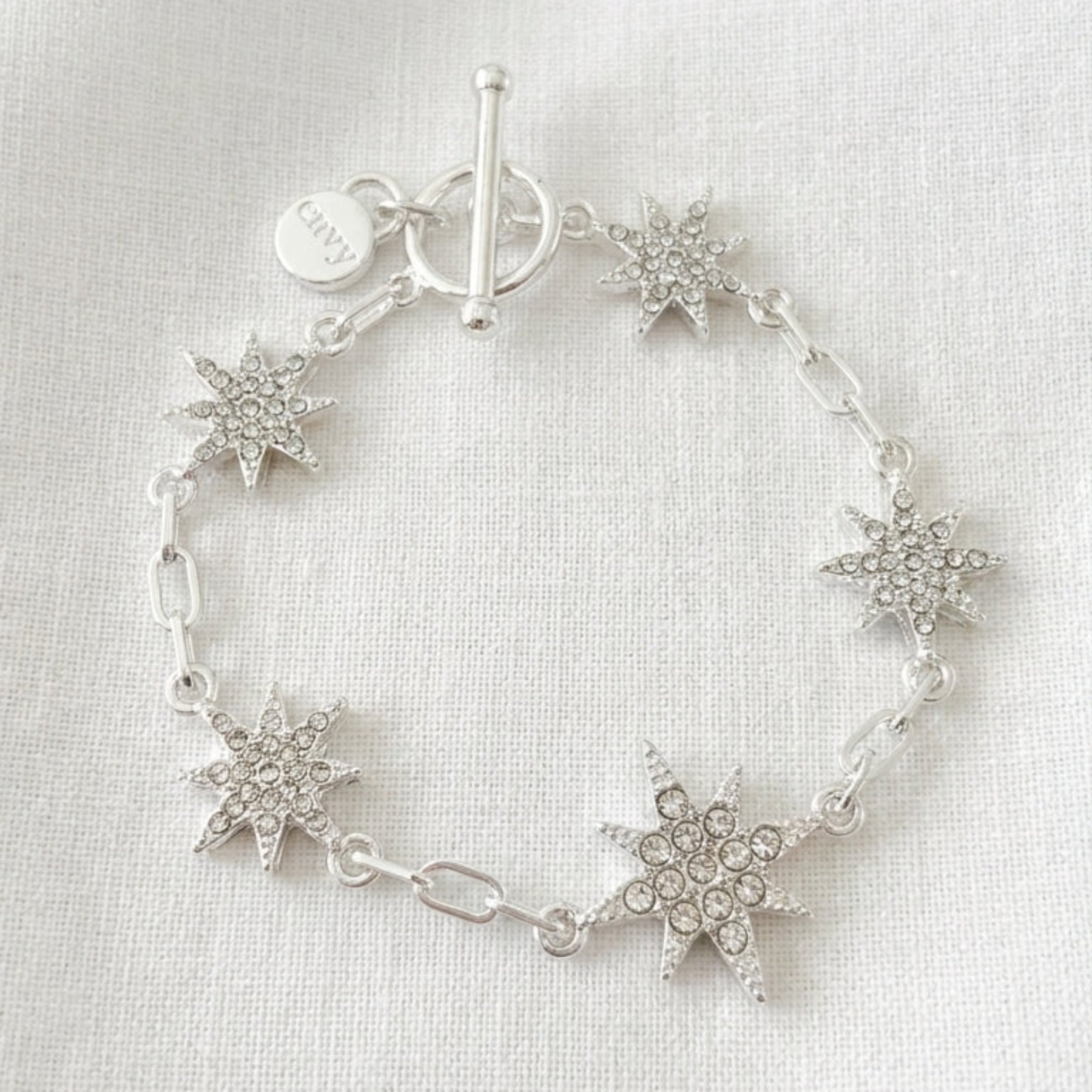Envy silver paperclip chain bracelet with six fully pavé crystal eight-pointed star charms and T-bar toggle clasp on cream linen background