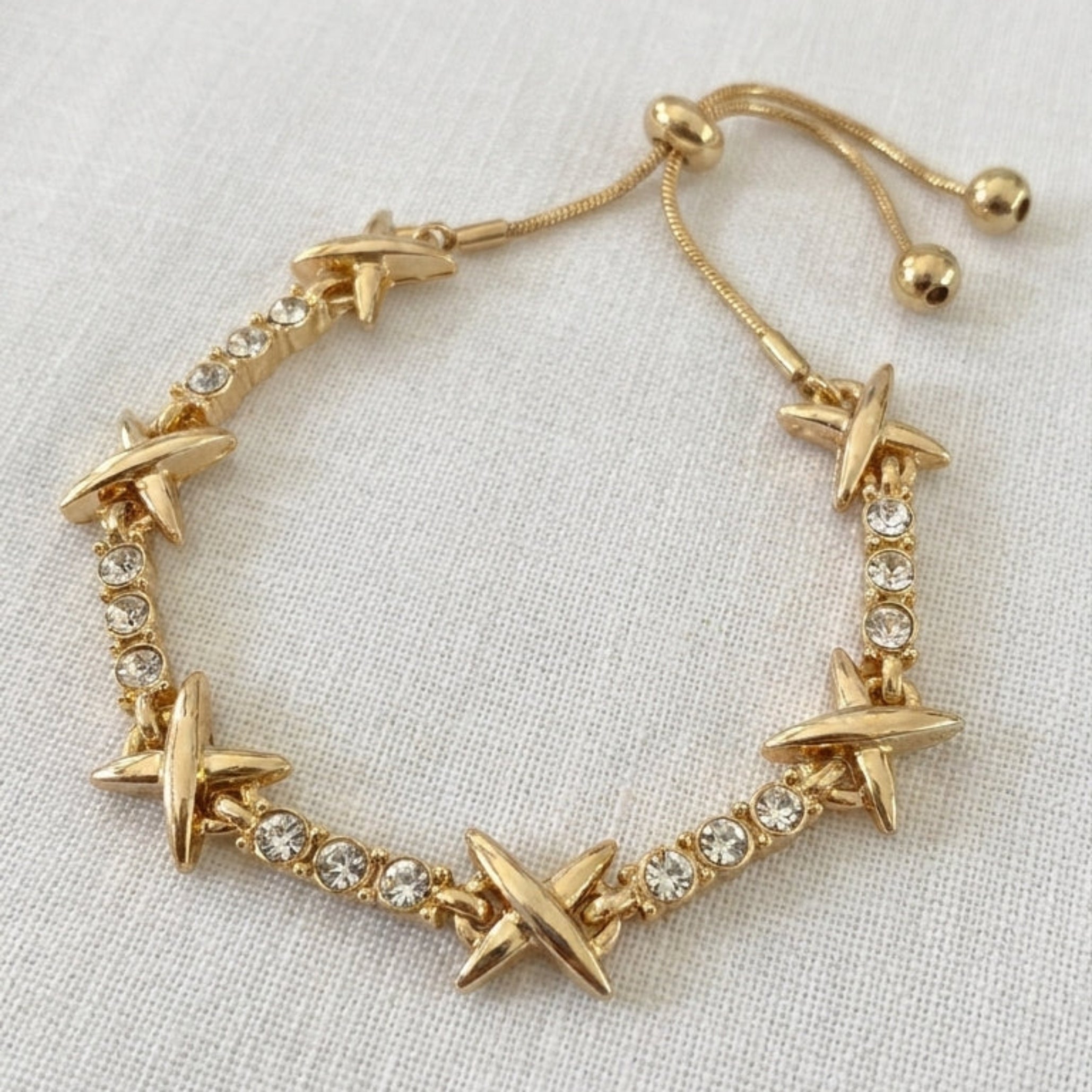 Envy gold adjustable slider bracelet alternating polished X-cross charms and round crystal links on cream linen background