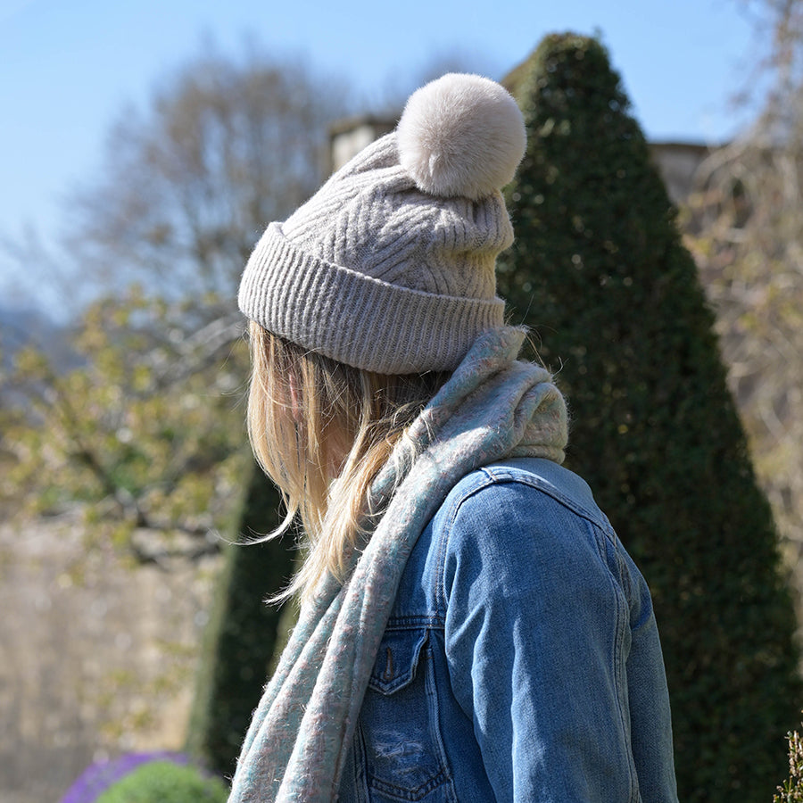 Side profile of model wearing the POM Charlotte oatmeal herringbone knit bobble hat with fluffy pom pom, styled with a denim jacket and grey scarf in a formal garden with topiary hedges on a sunny day.