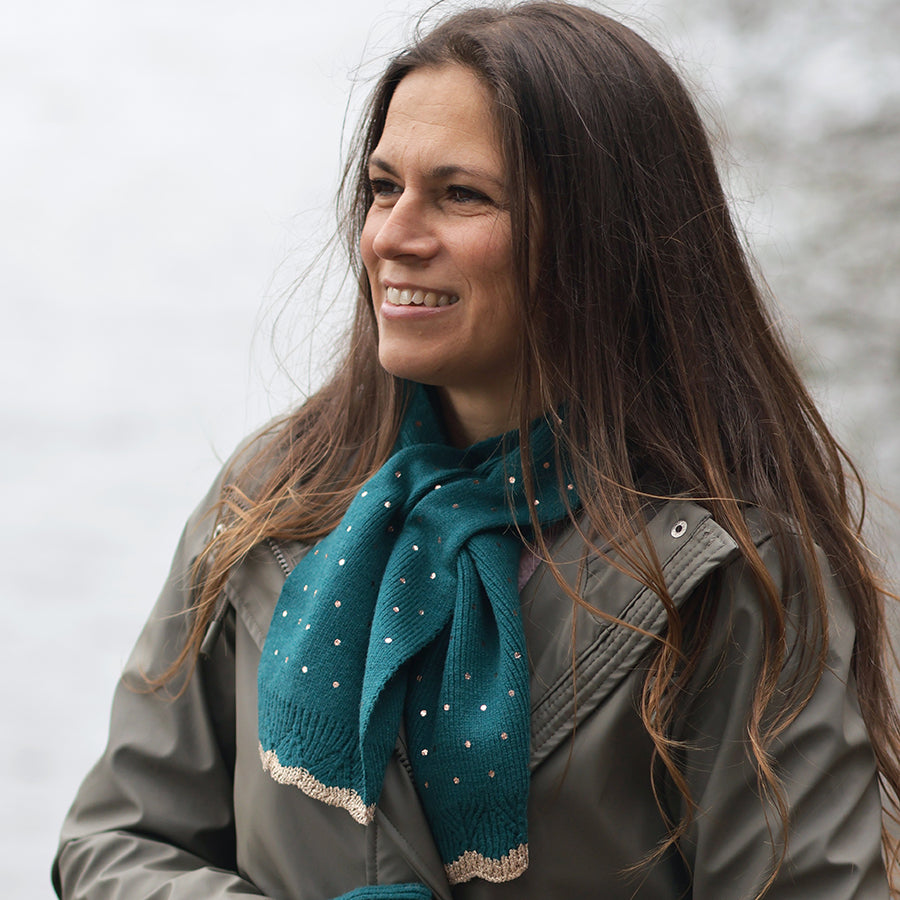 Smiling model looking to the side outdoors by a misty lake wearing the POM Yasmin teal metallic spot pull-through scarf loosely knotted at the neck, styled with a khaki waxed jacket and long wavy hair.