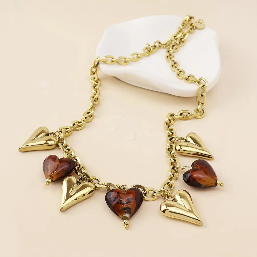 POM Treasure 14k gold plated stainless steel charm necklace featuring alternating polished gold and tortoiseshell brown heart pendants on a chunky chain, against a beige background."