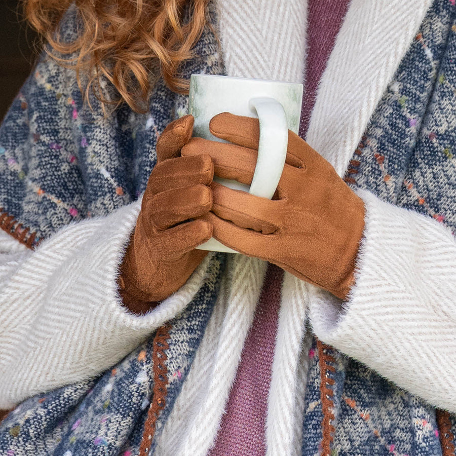 Model holding a ceramic mug with both hands wearing POM Kate tan faux suede gloves, styled with a multicolour knit wrap scarf and cream herringbone cardigan.