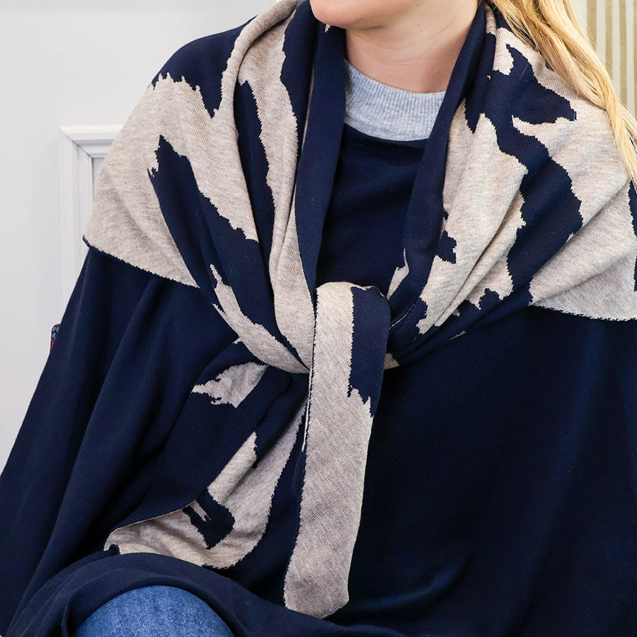 Model wearing the POM navy and oatmeal abstract animal print triangle scarf wrapped around the neck and draped over a navy poncho, styled with a grey knit jumper and jeans.
