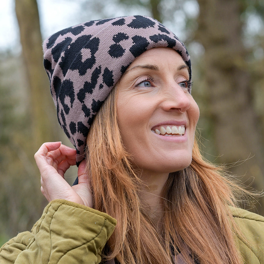 Smiling model looking upward wearing the POM Carly taupe and black leopard print beanie with a relaxed slouchy fit, styled with an olive green quilted jacket outdoors in a woodland setting.