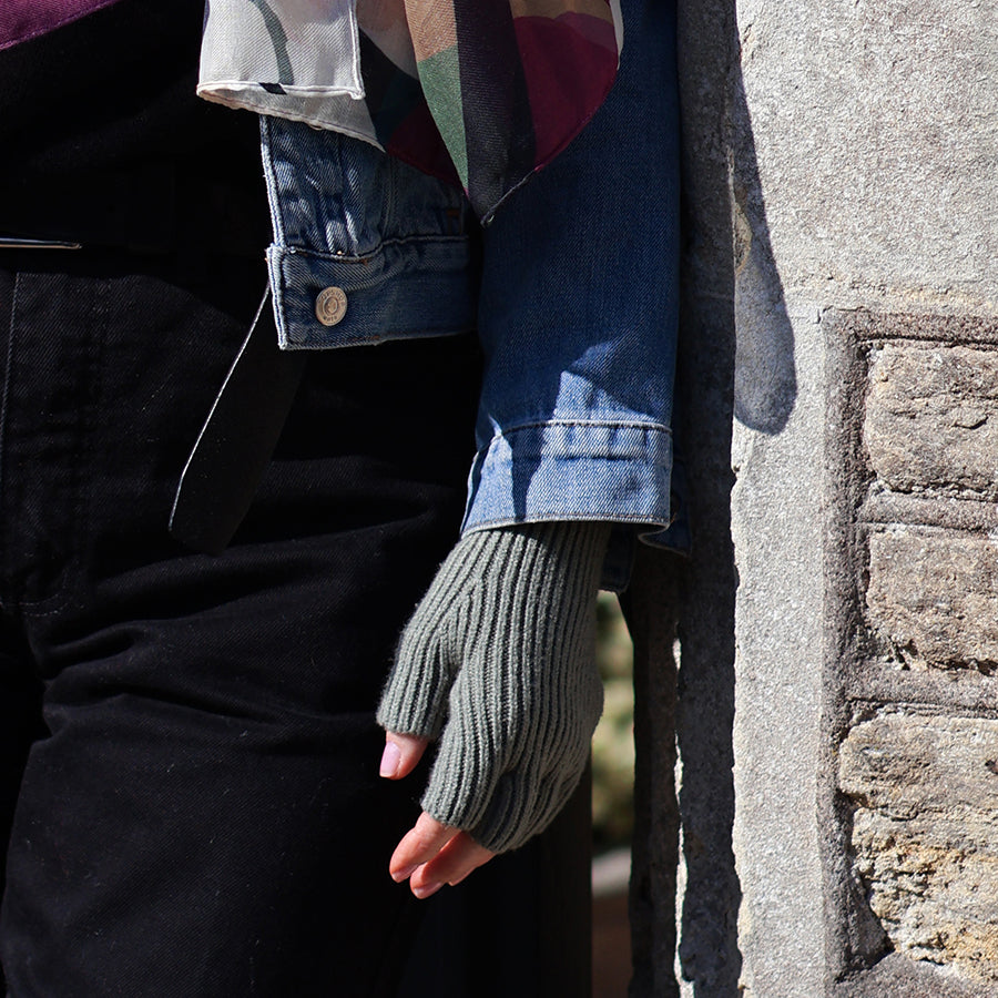Model wearing POM Yasmin sage green ribbed fingerless gloves, styled with a denim jacket and check scarf, hand resting against a stone wall outdoors.