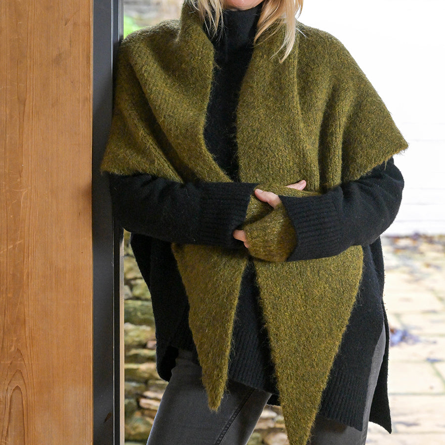 Model wearing the POM Ashleigh khaki chunky knit wrap scarf draped over both shoulders like a shawl, styled with a black turtleneck, black fingerless gloves and leather trousers, leaning against a wooden doorframe outdoors.