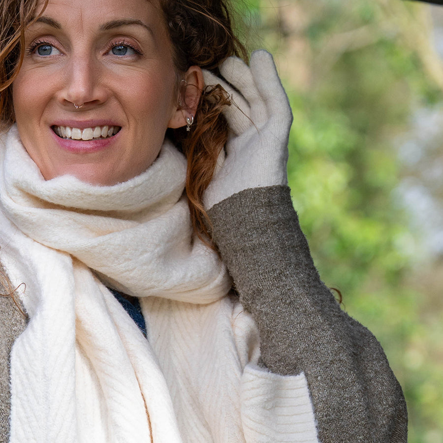 Smiling model tucking hair behind her ear wearing POM Charlotte cream cable knit gloves, styled with a matching cream herringbone scarf and a grey knit jumper outdoors with green foliage in the background.
