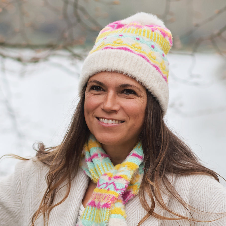 Smiling model outdoors in a snowy setting wearing the POM Mia cream and neon Fair Isle bobble hat with fluffy pom pom, styled with the matching Mia neon scarf and a cream textured coat.