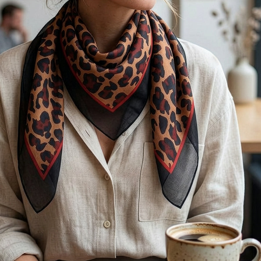 Model seated in a café holding a coffee cup wearing the POM camel and navy leopard print silk scarf loosely knotted at the neck, styled with a cream linen shirt.