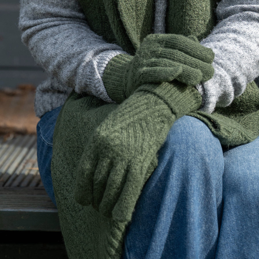 Model seated on a bench with hands resting on knees wearing POM Charlotte dark khaki cable knit gloves, styled with a dark khaki knit cardigan, grey jumper and blue jeans outdoors.