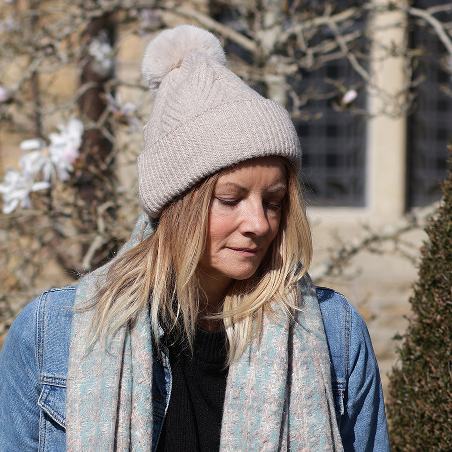 Model wearing the POM Charlotte oatmeal herringbone knit bobble hat with eyes downcast, styled with a denim jacket and a pale blue floral print scarf in a sunny garden setting with magnolia blossom.