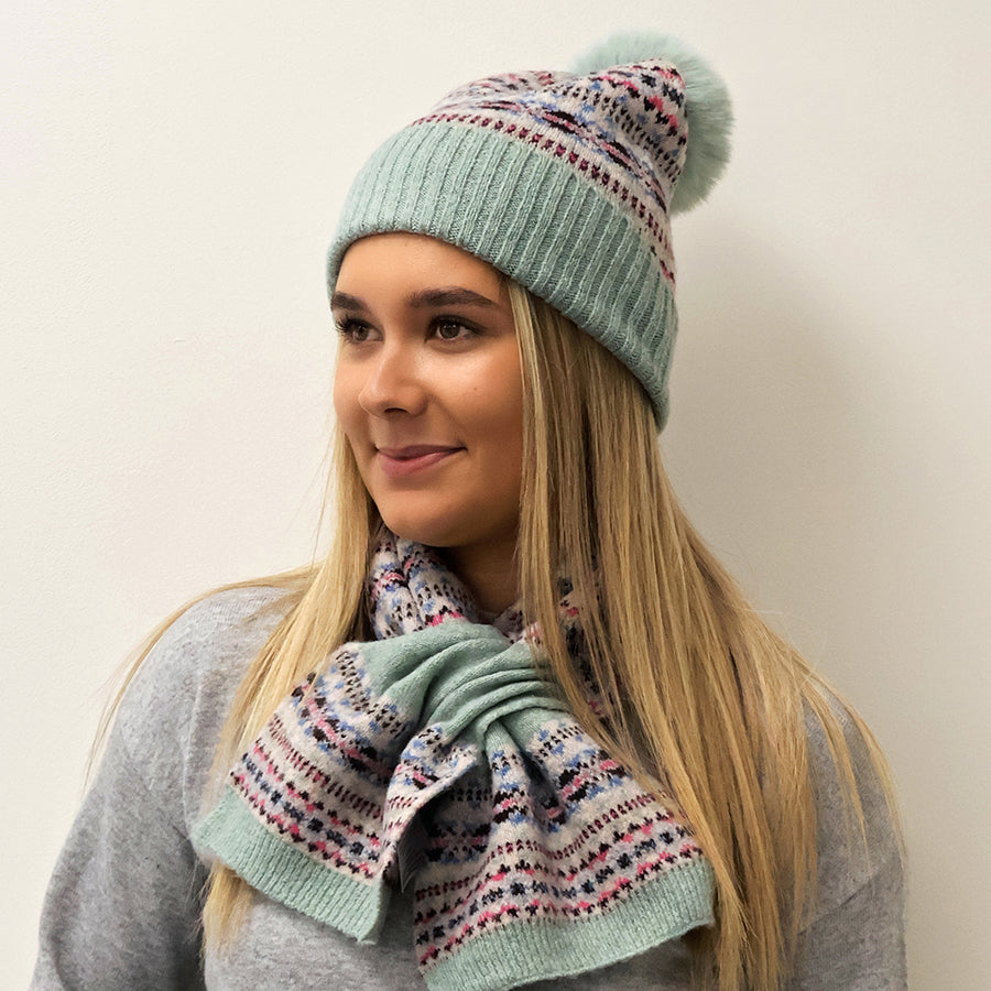 Model wearing the POM Iris pink and mint fair isle bobble hat with matching Iris fair isle scarf in mint trim, styled with a grey knit jumper against a white wall.