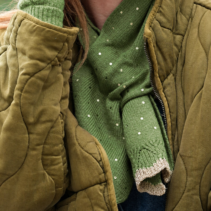 Close-up of model wearing the POM Yasmin green metallic spot pull-through scarf tucked into an olive quilted jacket, showing the sparkling sequin spots and gold lurex trim edge with a green bobble hat visible above.