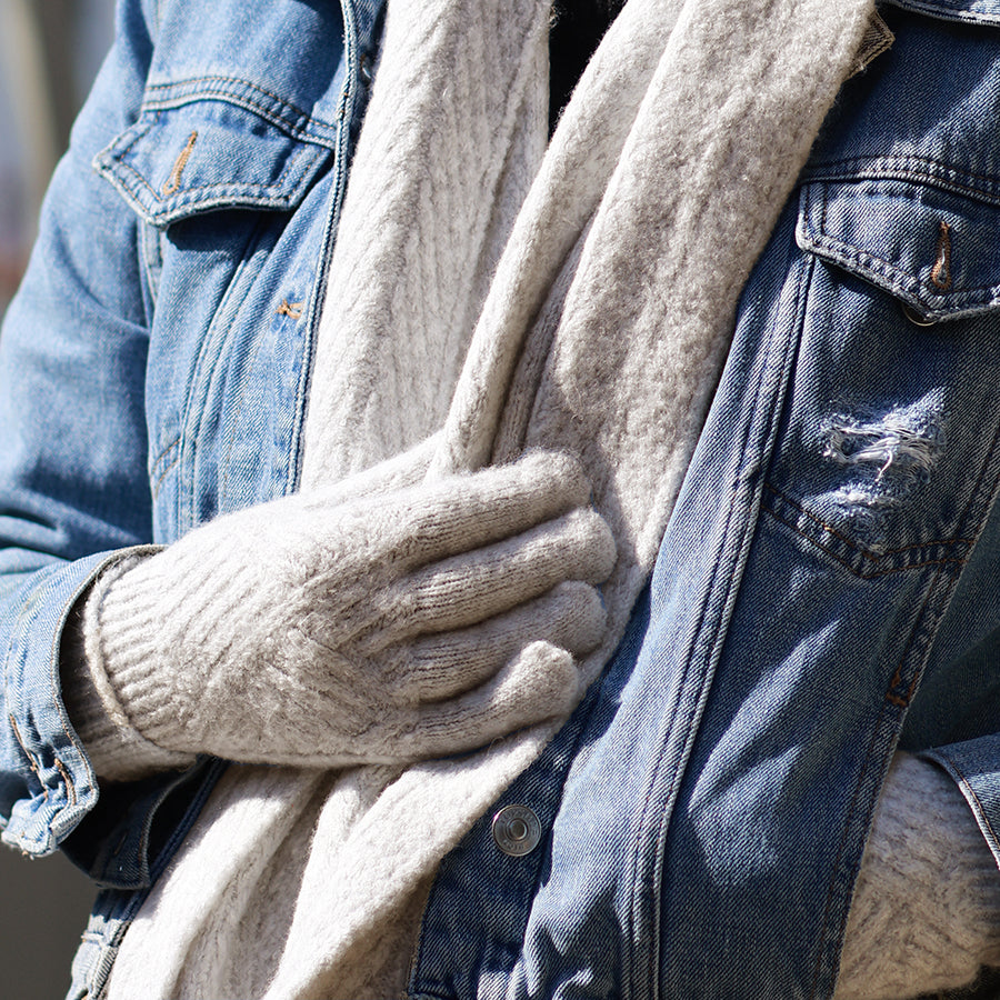 Model with arms crossed holding an oatmeal chunky knit scarf wearing POM Charlotte oatmeal cable knit gloves, styled with a denim jacket in bright winter sunlight outdoors.