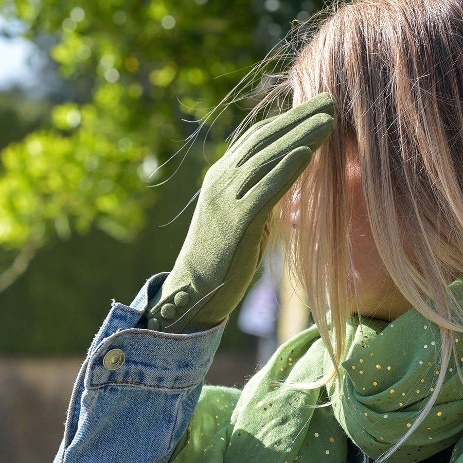 Model raising her hand to her hair outdoors wearing POM Kate sage green faux suede gloves, showing the three-button wrist detail, styled with a denim jacket and green spotted scarf with green foliage in the background.